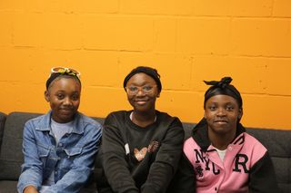 Three young people sitting on a grey sofa the wall behind them is orange 