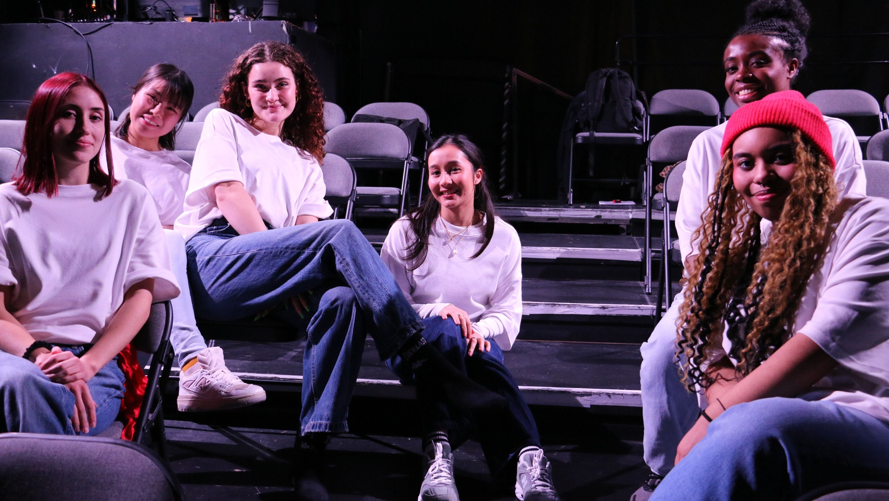 Hackney Young Collect Company A group of young women in white T-shirts smiling to the camera. They are sat in the audience seats at a theatre