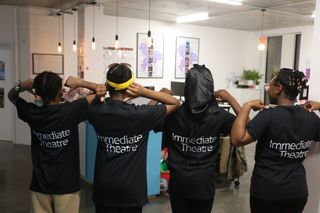 Four young people with their backs turned to the camera. They are all pointing to their back. On the back of their shirt is written 'Immediate Theatre'