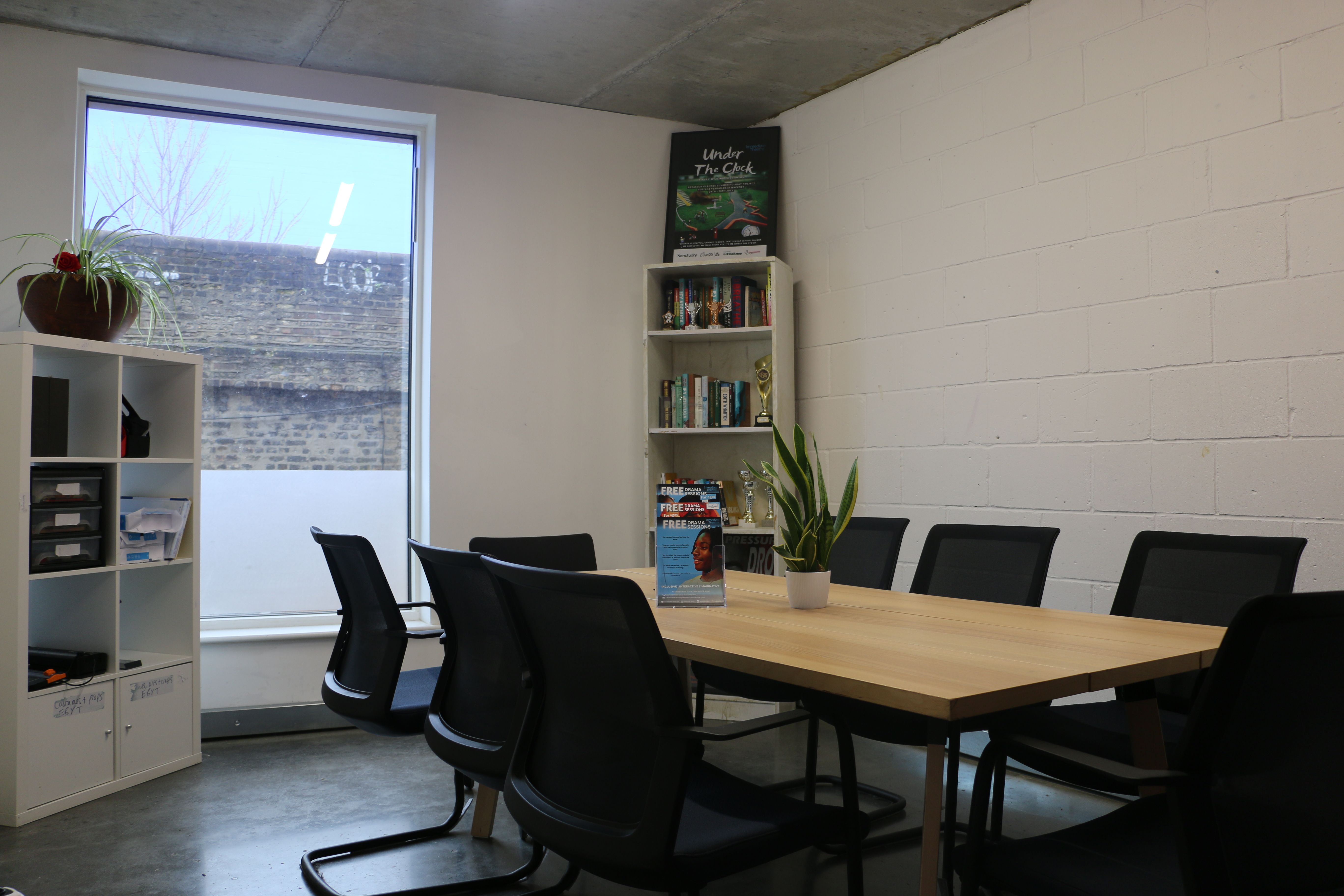 Room with a large table in the the middle. There are several chairs around the table and a large window at the rear of the room flagged by two book shelves on either side